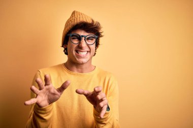 Genç yakışıklı bir adam günlük tişört ve gözlük takıyor sarı arka plandaki tiksindirici ifade, tiksindirici tepki verdiği için tiksindirici ve tiksindirici bir ifade takınıyor. Eller havada