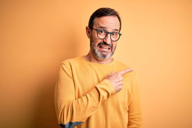 Orta yaşlı, beyaz bir adam, sarı arka planda gözlüklü, sıradan bir süveter giyiyor. Endişeli ve endişeli bir ifade takınıyor.