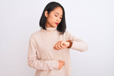 Balıkçı yaka kazak giyen genç bir kadın izole edilmiş beyaz arka planda duruyor. Saati kontrol ediyor, rahat ve kendinden emin.