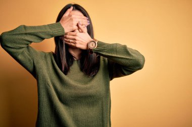 Mavi gözlü genç esmer kadın sarı arka planda yeşil süveter giyiyor gözleri ve ağzı kapalı, şaşırmış ve şok olmuş. Duygularını gizliyor.