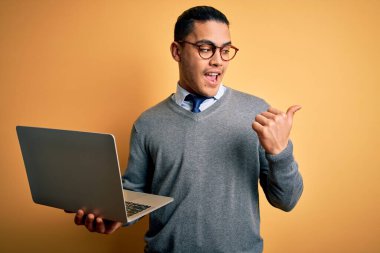 Genç Brezilyalı iş adamı, dizüstü bilgisayarını kullanarak sarı arka planda duruyor ve gülümseyen bir gülümsemeyle baş parmağıyla işaret ediyor.