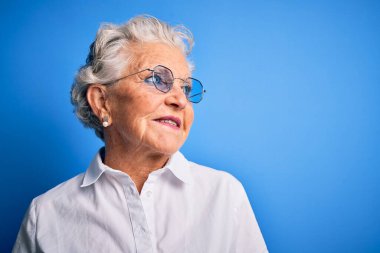 Zarif gömlek ve gözlük takan son sınıf güzeli bir kadın izole edilmiş mavi arka planda yüzünde gülümsemeyle, doğal bir ifadeyle yan yana bakıyor. Kendinden emin gülen.