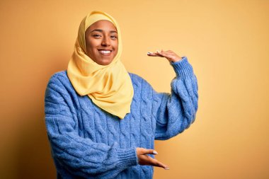 Genç Afrikalı Afro kadın, izole edilmiş sarı arka plan üzerine Müslüman başörtüsü takıyor. Ellerinde büyük ve büyük boy işareti, ölçü sembolü var. Kameraya bakıp gülümsüyor. Ölçüm konsepti.