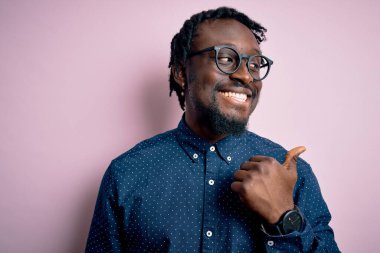 Genç, yakışıklı, Afro-Amerikalı bir adam pembe arka planda günlük gömlek ve gözlük takıyor. Gülümsüyor, gülümsüyor ve başparmağıyla yan tarafa işaret ediyor..