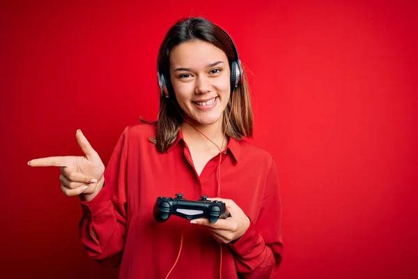 年轻漂亮的黑发女孩玩电子游戏时 用操纵杆和耳机 很高兴地将手和手指指向旁边 — 图库照片