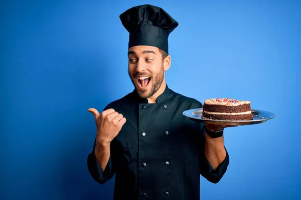 Young handsome cooker man with beard wearing uniform and hat holding tray with cake pointing and showing with thumb up to the side with happy face smiling