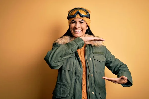 大小のサインを示す手で冬の天候ジェスチャーのためのスキーメガネとコートを身に着けている若い美しいヒスパニック系の女性は シンボルを測定します カメラを見て微笑んだ 概念の測定 — ストック写真