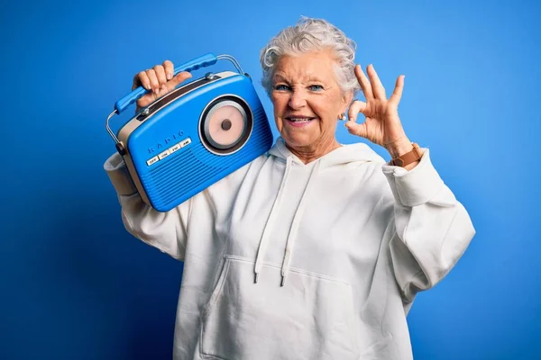 年长漂亮的女人拿着老式收音机 站在孤零零的蓝色背景上 手握手指 做着好的手势 很好的符号 — 图库照片