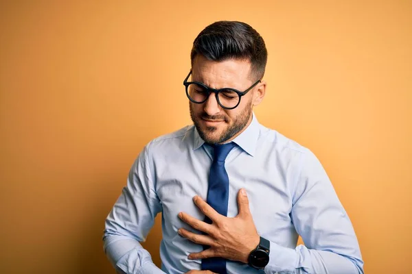 若いハンサムなビジネスマンのネクタイや眼鏡を胃の上に手で黄色の背景の上に立って消化不良 痛みを伴う病気の気分が悪い アチェの概念 — ストック写真
