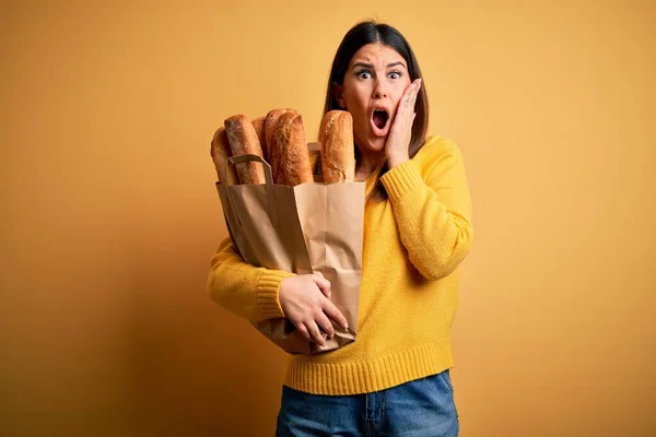 年轻美丽的女人 提着一袋黄色背景的健康的新鲜面包 脸上带着恐惧 惊奇和惊奇的表情 — 图库照片