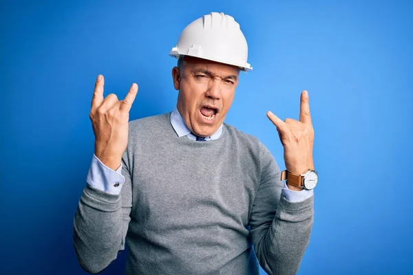 Middle age handsome grey-haired engineer man wearing safety helmet over blue background shouting with crazy expression doing rock symbol with hands up. Music star. Heavy concept.