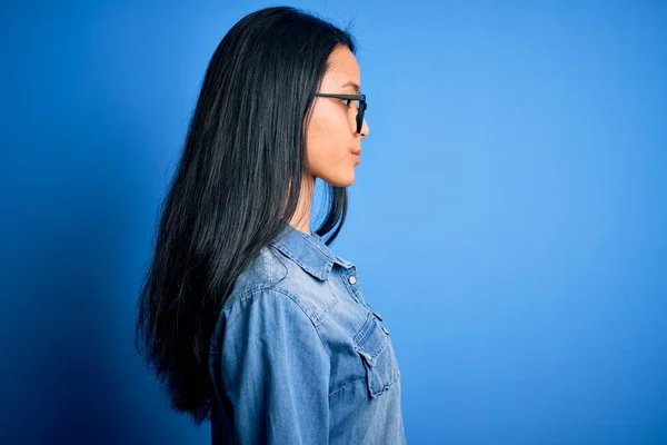 若いです美しいです中国の女性身に着けていますカジュアルデニムシャツ上の隔離された青の背景を探しています 自信を持って笑顔で自然な顔とリラックスしたプロフィールポーズ — ストック写真