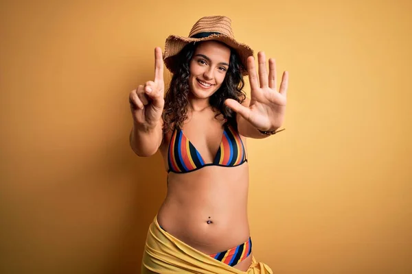 Young beautiful woman with curly hair on vacation wearing bikini and summer hat showing and pointing up with fingers number six while smiling confident and happy.