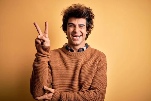 カジュアルなシャツとセーターを身に着けている若いハンサムな男は カメラでの勝利サインを指で行う幸せな顔のウィンクで笑顔で孤立した黄色の背景に 第二番 — ストック写真