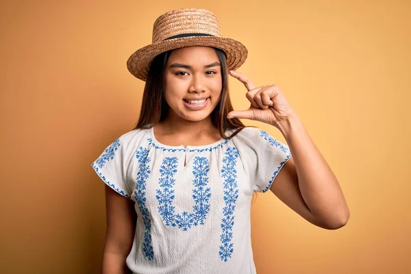 年轻美丽的亚洲女孩 身穿休闲装 头戴帽子 站在黄色的背景上 微笑着 自信地做手势 手拿着小尺寸的标志 手拿着手指 手拿着相机 计量概念 — 图库照片