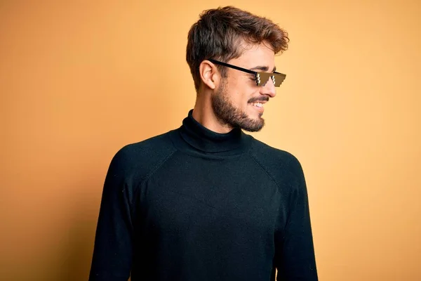 Young man wearing thug life fanny sunglasses standing over isolated yellow background looking away to side with smile on face, natural expression. Laughing confident.