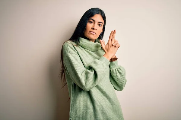 Young beautiful hispanic woman wearing green winter sweater over isolated background Holding symbolic gun with hand gesture, playing killing shooting weapons, angry face
