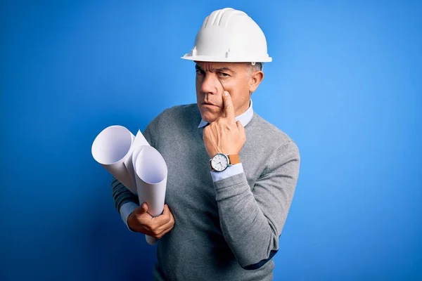 Middle age handsome grey-haired architect man wearing safety helmet holding blueprints Pointing to the eye watching you gesture, suspicious expression