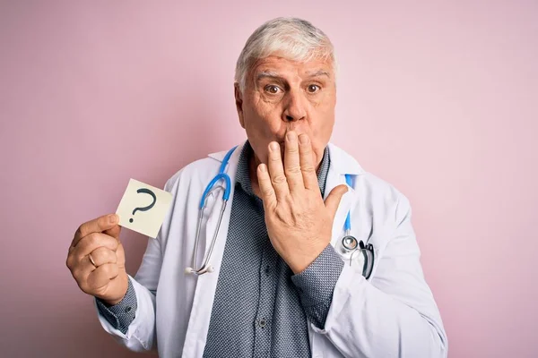 Senior handsome hoary doctor man wearing stethoscope holding reminder with question mark cover mouth with hand shocked with shame for mistake, expression of fear, scared in silence, secret concept