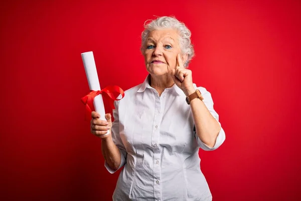 Senior beautiful student woman holding diploma standing over isolated red background serious face thinking about question, very confused idea