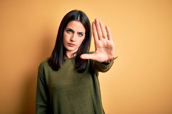 Young brunette woman with blue eyes wearing green casual sweater over yellow background doing stop sing with palm of the hand. Warning expression with negative and serious gesture on the face.