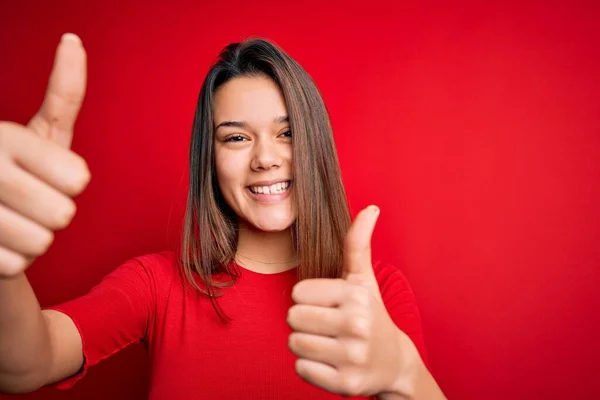Young beautiful brunette girl wearing casual t-shirt over isolated red background approving doing positive gesture with hand, thumbs up smiling and happy for success. Winner gesture.