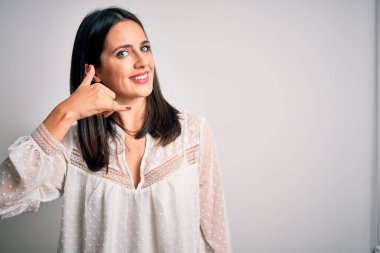 Mavi gözlü genç esmer kadın izole edilmiş beyaz arka plan üzerine günlük tişört giyip telefonla el ve parmak hareketleriyle gülümsüyor telefonda konuşur gibi. İletişim kavramları.