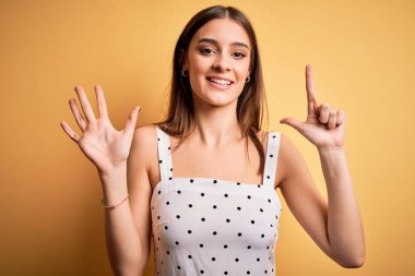 Sarı arka planda dikilen, rahat elbiseler giyen genç, güzel esmer kadın yedinci parmaklarıyla işaret ederken kendinden emin ve mutlu bir şekilde gülümsüyor..