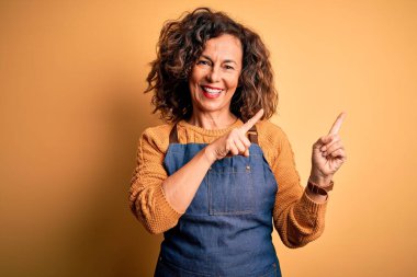 Orta yaşlı, güzel fırıncı kadın, önlük giymiş, izole edilmiş sarı arka planda duruyor gülümsüyor ve kameraya bakıyor, iki eliyle ve parmaklarıyla işaret ediyor..