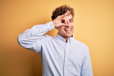 Sarışın, kıvırcık saçlı, sarı arka planda çizgili tişört giyen, el hareketleriyle gülümseyen, mutlu yüzüyle parmaklarının arasından bakan genç, yakışıklı bir adam..