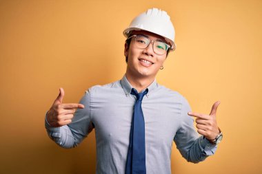 Güvenlik kaskı takan ve sarı arka planda kravat takan genç yakışıklı mimar yüzünde gülümsemeyle gururlu ve mutlu birini işaret ediyor..