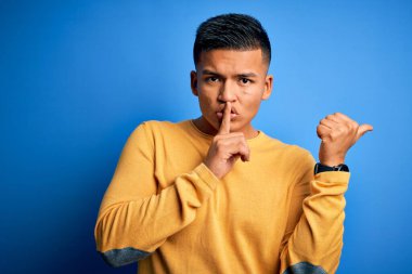 İzole edilmiş mavi arka planda sarı günlük süveter giyen genç, yakışıklı latin adam ellerini yana uzatarak parmaklarını dudaklarına uzatarak sessiz olmak istiyor. Sessizlik ve gizli konsept.