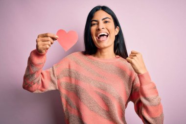 Genç ve güzel İspanyol bir kadın pembe arka planda romantik bir kalp kağıdı tutuyor. Gururla bağırıyor, zaferi kutluyor ve başarıyı kutluyor. Çok heyecanlı, coşkulu bir duygu.