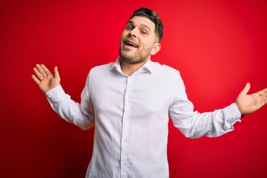 Mavi gözlü, zarif gömlekli genç bir iş adamı kırmızı izole edilmiş arka plan üzerinde duruyor. Kolları ve elleri havada, kafası karışmış bir yüz ifadesi var. Şüphe kavramı.