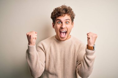 Young blond handsome man with curly hair wearing casual sweater over white background celebrating surprised and amazed for success with arms raised and open eyes. Winner concept.