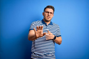 Mavi gözlü, gözlüklü ve mavi arkaplanlı sıradan çizgili tişörtlü, tiksindirici, tiksindirici, tiksindirici bir yüz ifadesi takınan genç adam. Eller havada