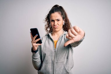 Kıvırcık saçlı güzel sporcu kız akıllı telefon ve kulaklık kullanarak müzik dinliyor. Kızgın yüzlü, negatif işaretli, baş parmakları aşağıda, reddedilme konsepti var.