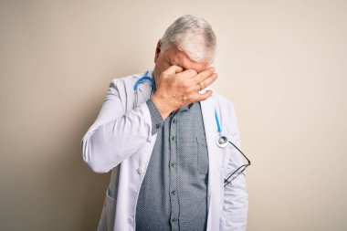 Beyaz arka plan üzerinde steteskop ve palto giyen kıdemli yakışıklı doktor yorgun burnu ve gözleri ovuşturup yorgun ve baş ağrısı çekiyor. Stres ve hüsran kavramı.