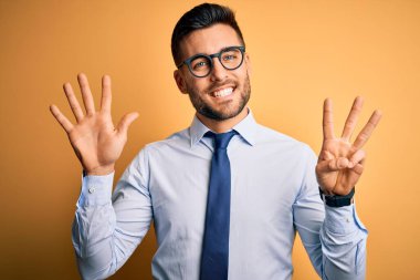Sarı arka plan üzerinde duran kravat ve gözlük takan genç yakışıklı işadamı sekiz numaralı parmaklarıyla gülümseyerek kendinden emin ve mutlu bir şekilde işaret ediyor..