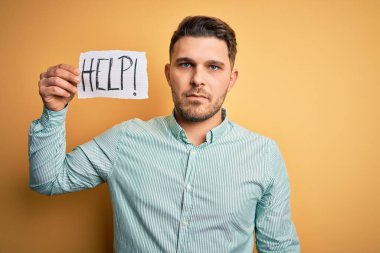 Mavi gözlü genç bir iş adamı sarı arka planda kağıt tutarken yardım istiyor. Zeki bir yüz ifadesiyle ciddi ciddi düşünüyor.