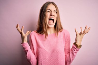 Genç, güzel, kızıl saçlı bir kadın pembe arka plan üzerine günlük süveter giyiyor çılgın ve sinirli bir şekilde bağırıyor ve agresif bir ifade ve kollarla bağırıyor. Hayal kırıklığı konsepti.