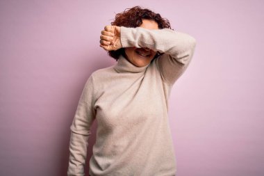 Orta yaşlı güzel kıvırcık saçlı kadın pembe arka plan üzerine balıkçı yaka kazak giyip gözleri kapayarak neşeli ve komik gülümsüyor. Kör kavram.