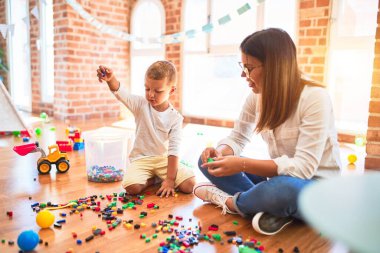 Güzel öğretmen ve bebek anaokulunda bir sürü oyuncağın etrafında yapı taşlarıyla oynuyorlar.