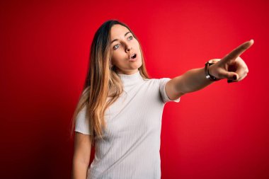 Güzel sarışın kadın, mavi gözlü, kırmızı arkaplan üzerine gündelik beyaz tişört giyen, parmağı önde olan, ağzı açık, şaşırmış, ön tarafta bir şey var.