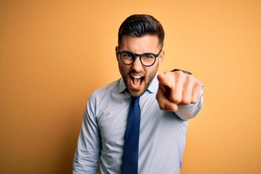 Sarı arka plan üzerinde duran kravat ve gözlük takan genç yakışıklı işadamı kameraya kızgın ve sinirli bir şekilde bakıyor.