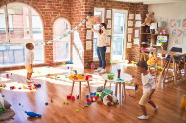 Anaokulunda bir sürü oyuncağın etrafında top ve hasır sepet kullanarak basketbol oynayan güzel öğretmenler ve yeni yürümeye başlayan çocuklar.
