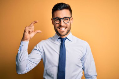 Sarı arka plan üzerinde duran kravat ve gözlük takan genç yakışıklı işadamı gülümsüyor ve kendine güvenen el hareketleriyle küçük beden işareti yapıyor ve kameraya bakıyor. Ölçü kavramı.