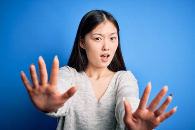 Genç, güzel Asyalı bir kadın, günlük süveter giyer, mavi izole arka planda durur, avuç içi hareketlerini durdurur, sinirli ve hüsran dolu bir ifade takınır.