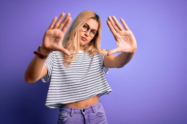 Genç, sarışın, çizgili tişört ve gözlük takan, mor arka planda el ve parmakları kullanarak çerçeve yapan güzel bir kadın.