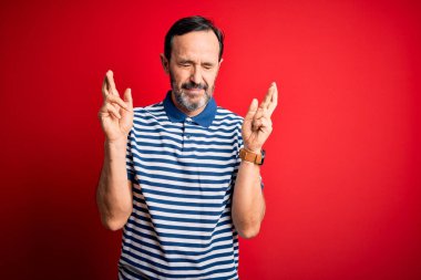 Orta yaşlı, beyaz, sıradan çizgili polo giyen, izole edilmiş kırmızı arka plan üzerinde duran umut ve gözler kapalı bir şekilde gülümseyerek el hareketi yapan bir adam. Şans ve batıl inanç..
